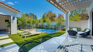 A modern outdoor patio with a glass table, two wicker chairs, a pool, and a pergola.