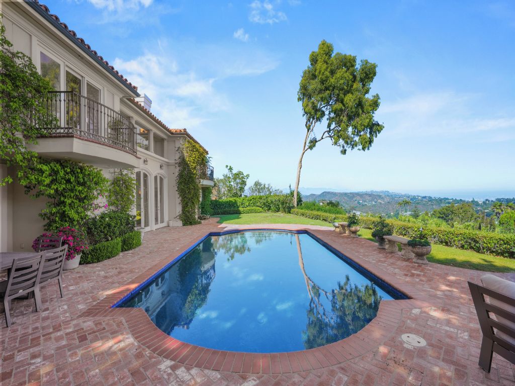 A swimming pool with a view of the city, surrounded by a brick patio and a garden.