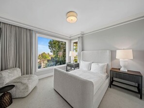 A modern hotel room with a large bed, a sitting area with a sofa, a nightstand with lamps, and a view of greenery outside.