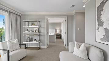 A modern hotel room with a neutral color palette, a large window, a sofa, a bookshelf, and a painting on the wall.