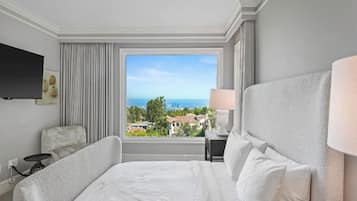 Ein Hotelzimmer mit einem großen Bett, einem Fernseher, einer Lampe und Blick auf Stadt und Meer.