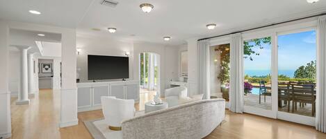 Amplia sala de estar con un televisor de pantalla plana grande, muebles blancos y vistas a una piscina y jardín a través de puertas de cristal.