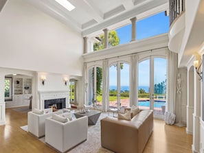 A spacious living room with a fireplace, large windows, and a view of a pool and greenery. - Savona Sanctuary Estate (Los Angeles)