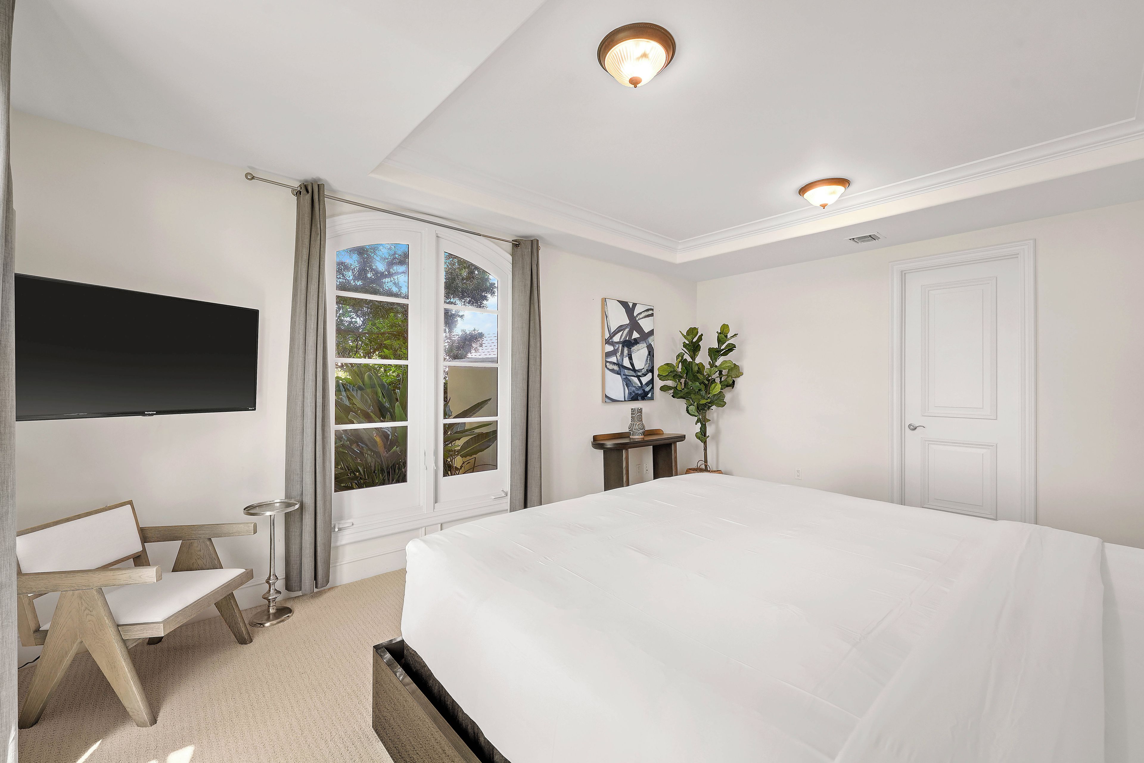 A bedroom with a large bed, a flat-screen TV mounted on the wall, a bedside table, and a potted plant.