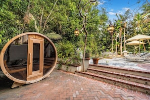 A circular wooden structure with a glass door, surrounded by lush greenery and a brick pathway leading to a seating area with umbrellas. - Sunset Vale (West Hollywood)