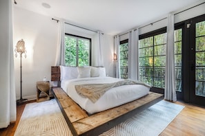 A spacious bedroom with a large bed, a wooden platform, and multiple windows offering a view of lush greenery. - Sunset Vale (West Hollywood)