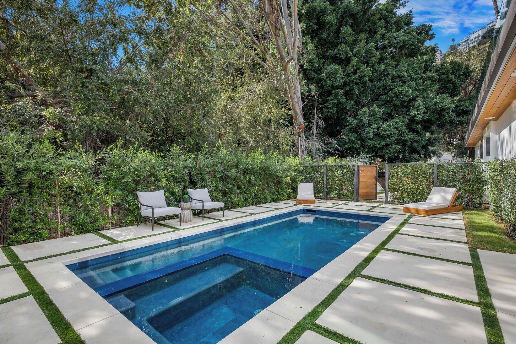 A rectangular swimming pool with a clear blue water surface, surrounded by a concrete deck with grass edging. There are two white lounge chairs and a wooden bench with a cushion. The pool area is enclosed by a wooden fence and dense green foliage.