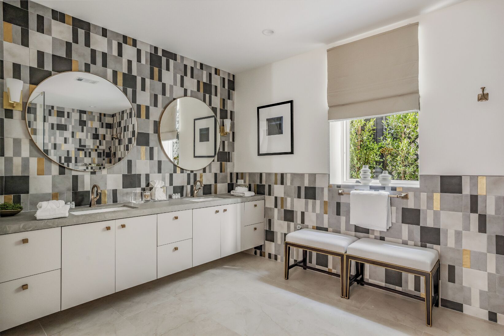 A modern bathroom with a checkered tile backsplash, a round mirror, a bench, and a window with a view of greenery.