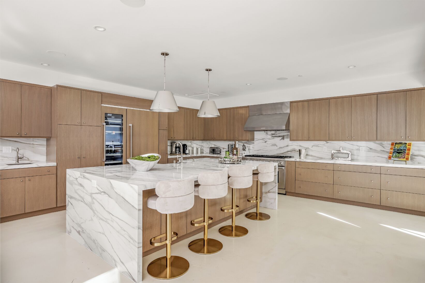 A modern kitchen with marble countertops, wooden cabinets, a central island with bar stools, and pendant lighting.