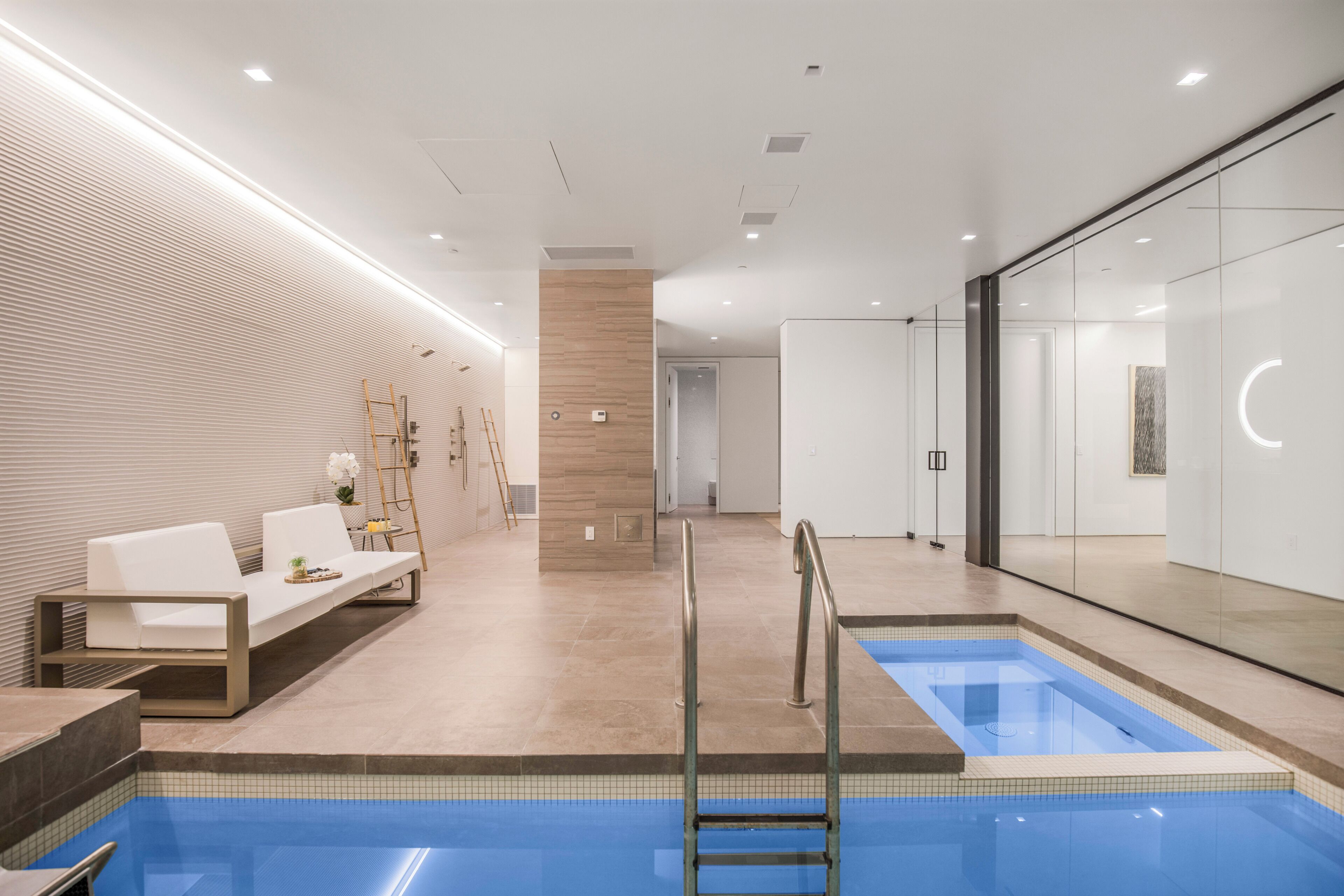 A modern indoor pool area with a lounge chair, a small table with a plant, and a pool with a ladder.