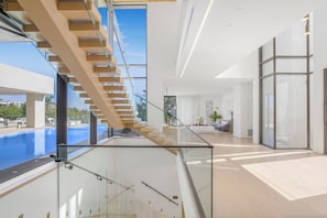 A modern interior with a glass staircase leading to a balcony overlooking a pool. - Swallow Sanctuary (West Hollywood)