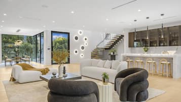 Un salon moderne avec un canapé, des fauteuils et une table basse. Un escalier mène à la cuisine.