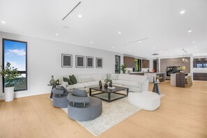 A modern living room with a white sofa, grey ottoman, and a coffee table. A kitchen is visible in the background. - Levitt Lane (Sherman Oaks)