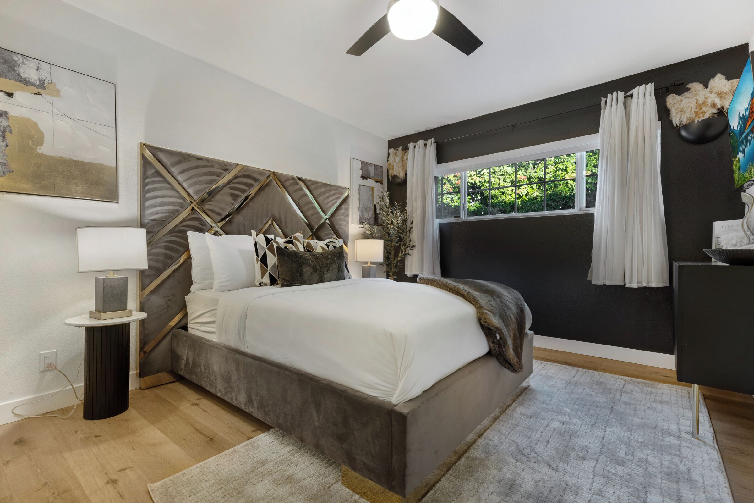 A modern bedroom with a large bed, a ceiling fan, a window with curtains, and a wall-mounted artwork.