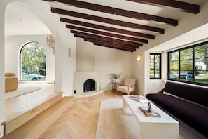 A modern living room with a fireplace, wooden beams, a sofa, and a view through large windows. - Bella Villa (Los Angeles)