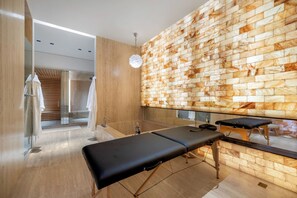 A modern spa room with a wooden floor, a massage table, a large mirror, and a stone wall. - Collingwood (West Hollywood)