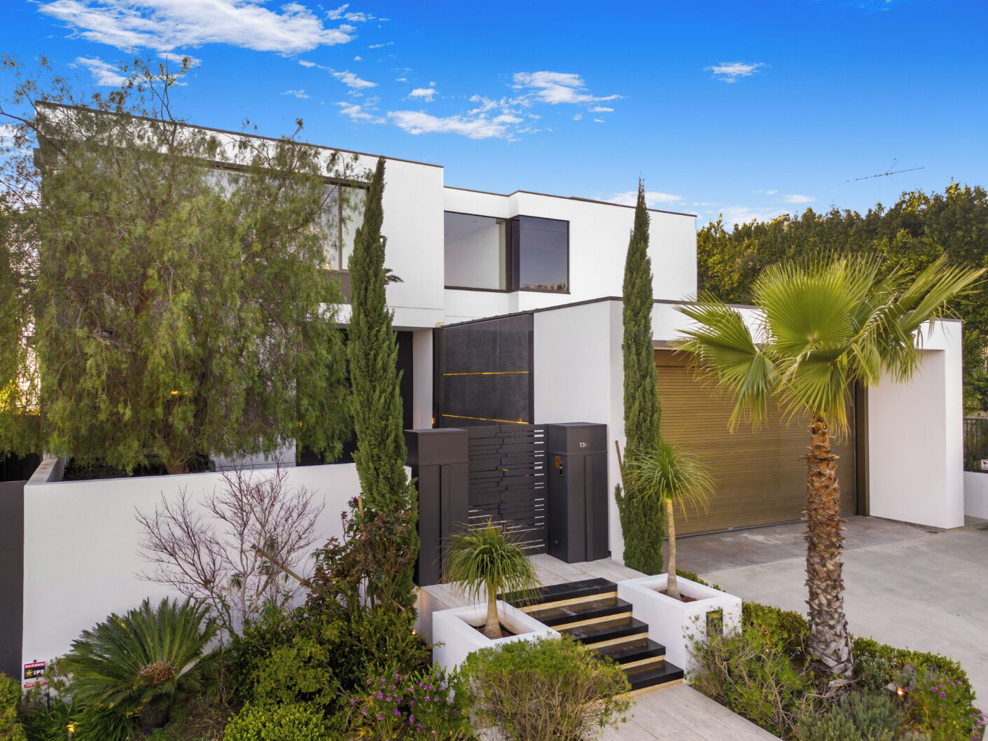 Una casa moderna de dos pisos con techo plano, garaje y un jardín bien cuidado con diversas plantas y una palmera.