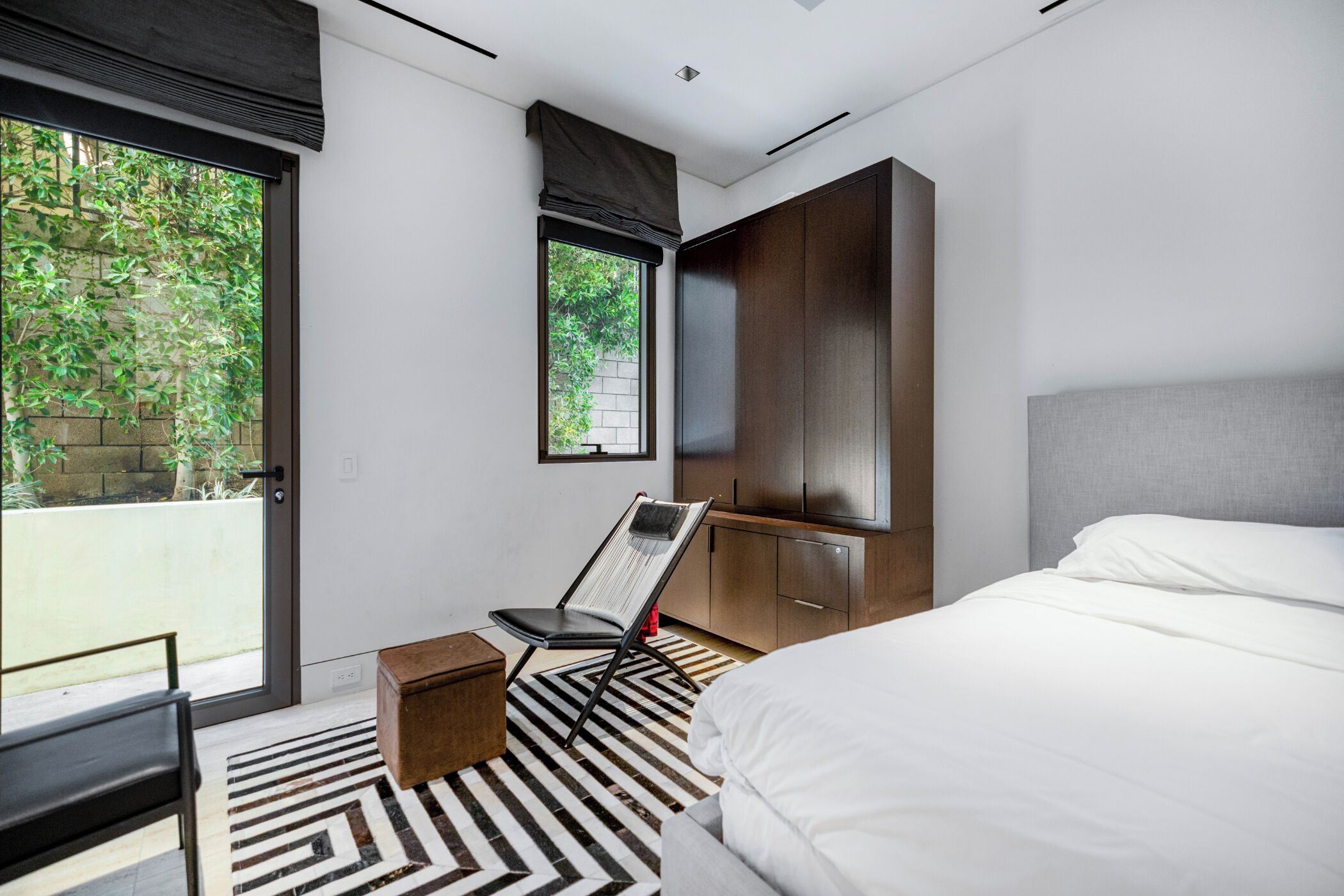 A modern bedroom with a bed, a chair, a small table, a wardrobe, and a window with a view of greenery.