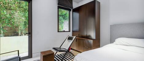 A modern bedroom with a bed, a chair, a small table, a wardrobe, and a window with a view of greenery.