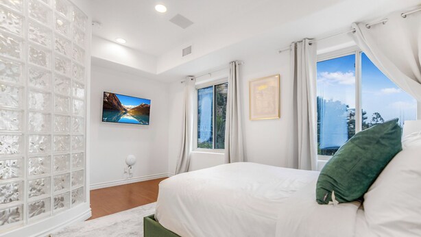 A bedroom with a large bed, a window with curtains, a wall-mounted TV, and a decorative glass partition. - Franklin Mediterranean Villa (West Hollywood)