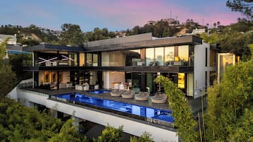 A modern two-story house with a large outdoor swimming pool, multiple seating areas, and a view of the surrounding landscape.