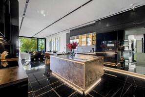 A modern kitchen with a marble island, black tiled floor, and a large window overlooking greenery. - London Dream (West Hollywood)