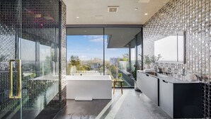 A modern bathroom with a glass-enclosed shower, a freestanding bathtub, and a large window offering a city view. - London Dream (West Hollywood)