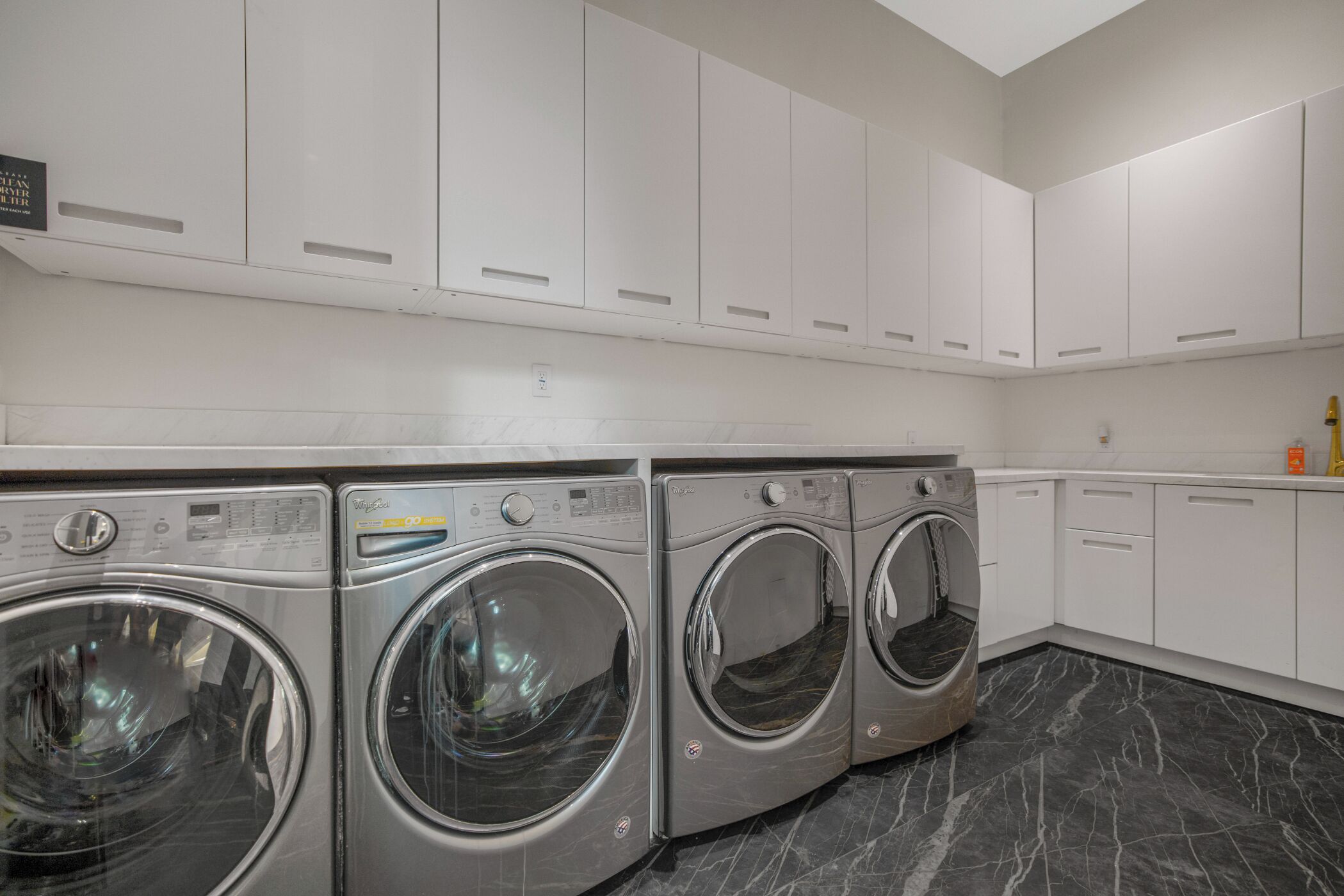 A laundry room with three front-loading washing machines, white cabinets, and a marble floor.