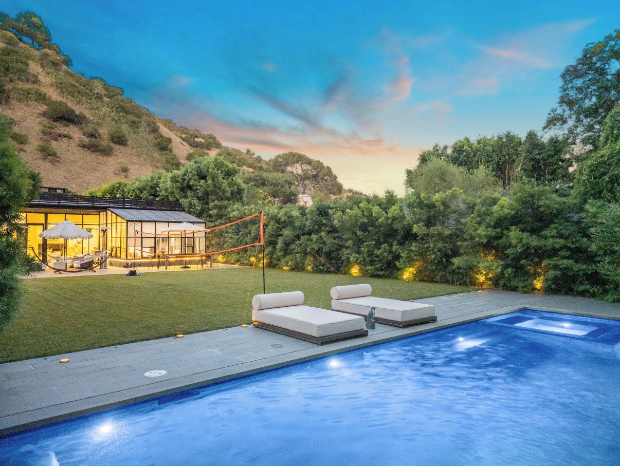 A swimming pool with lounge chairs, a glass-enclosed patio with outdoor furniture, and a hillside backdrop.