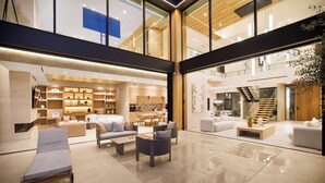 A spacious lobby with a modern design, featuring a seating area with sofas and chairs, a dining area, and a staircase leading to an upper level. - Hutton Villa (Beverly Hills)