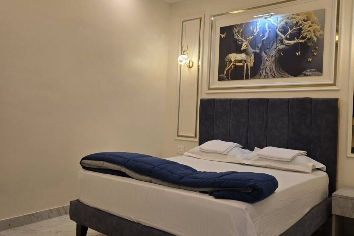 Una cama bien hecha con un cabecero oscuro, una obra de arte en la pared con un ciervo y un árbol, y una lámpara de pared.
