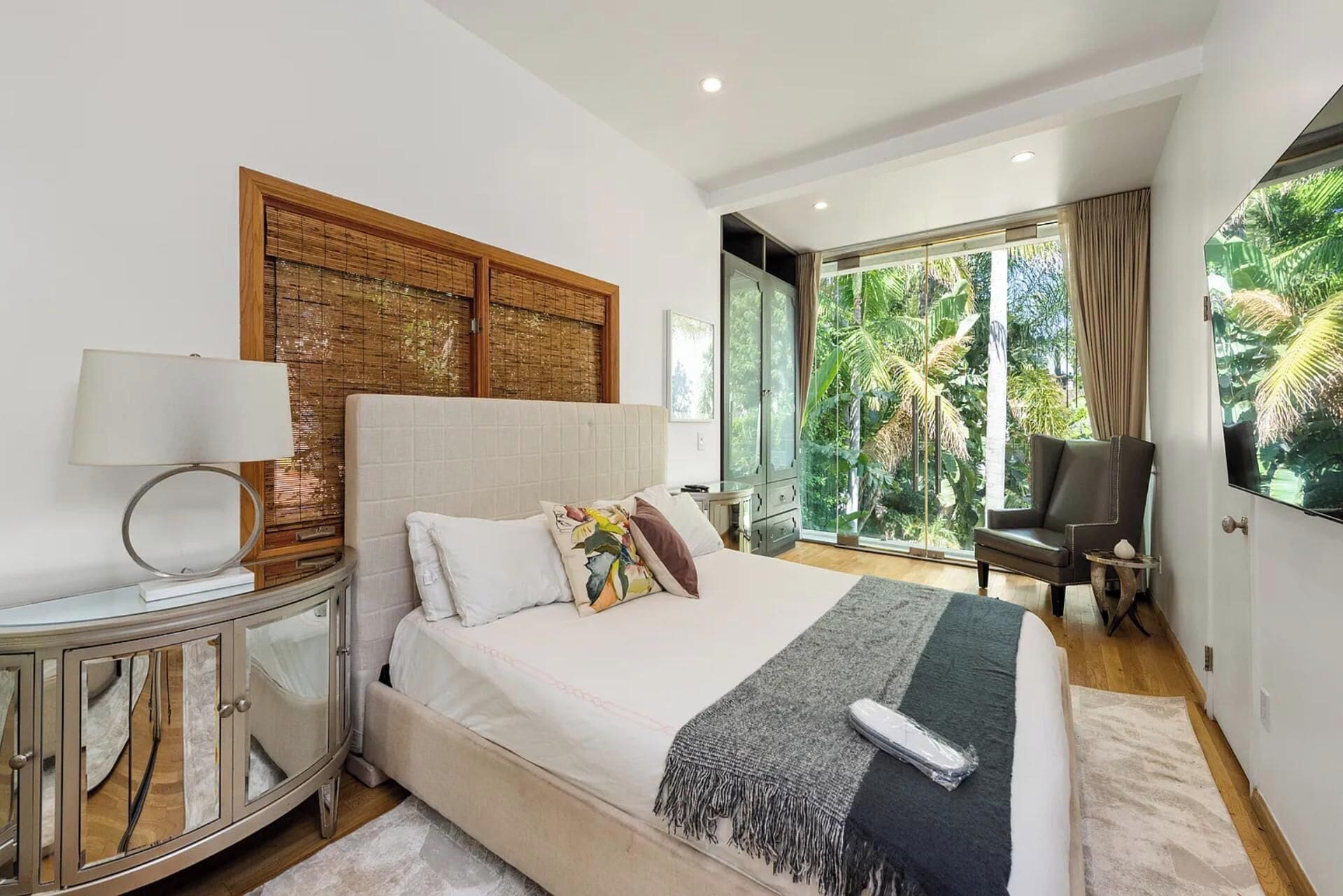 A modern bedroom with a large bed, a bedside table, a lamp, a chair, and a view of greenery through large windows.