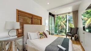 Un dormitorio moderno con una cama grande, una mesita de noche, una lámpara, una silla y vistas a zonas verdes a través de amplias ventanas.