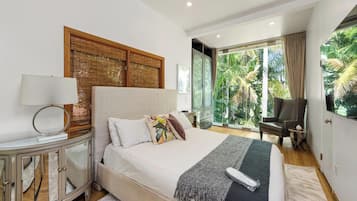 A modern bedroom with a large bed, a bedside table, a lamp, a chair, and a view of greenery through large windows.