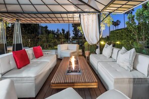 Una terrazza esterna con divani bianchi, un tavolo centrale in legno con elemento decorativo a fuoco e un tettoia sopra.