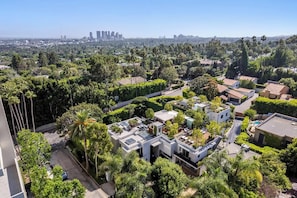 An aerial view of a residential area with modern houses, lush greenery, and a distant city skyline. - Alta Royale (West Hollywood)