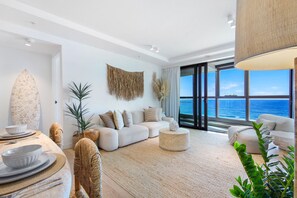 A modern living room with a large window overlooking the sea, a white sofa, a dining area with a round table, and a decorative wall piece. - Elegance Oceanview 2BR Suite -The Langham +Parking (Surfers Paradise)