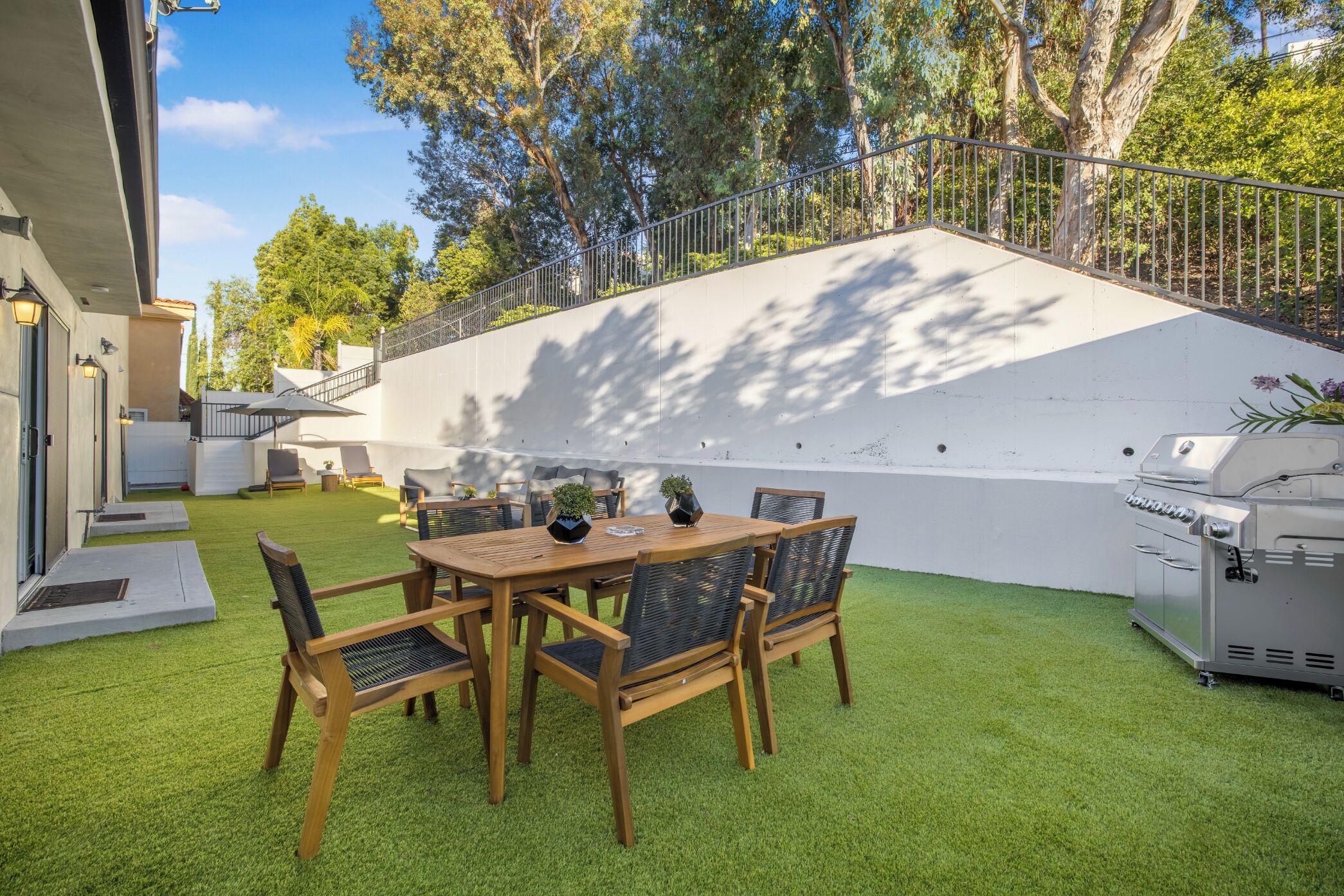 Un patio con mesa y sillas de madera, una parrilla y una pared blanca con valla metálica.