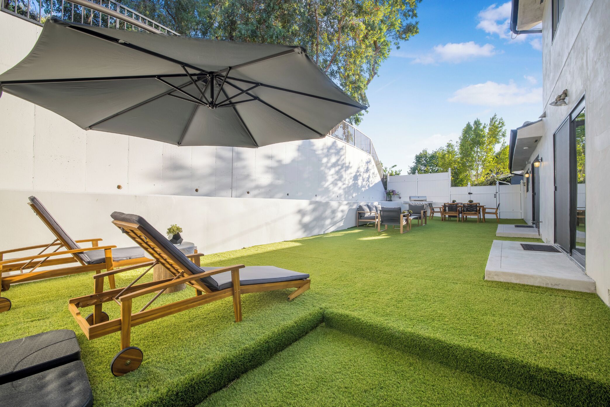 Un jardin avec une pelouse artificielle, des fauteuils de jardin en bois et un grand parasol.