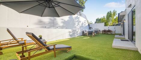 Un jardin avec une pelouse artificielle, des fauteuils de jardin en bois et un grand parasol.
