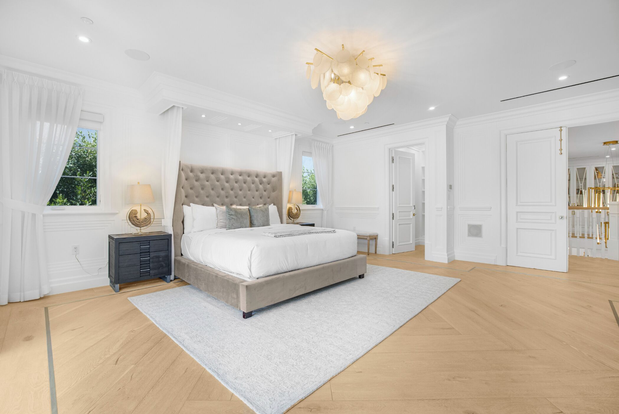 A spacious bedroom with a large bed, a nightstand, a chandelier, and a view of greenery outside.
