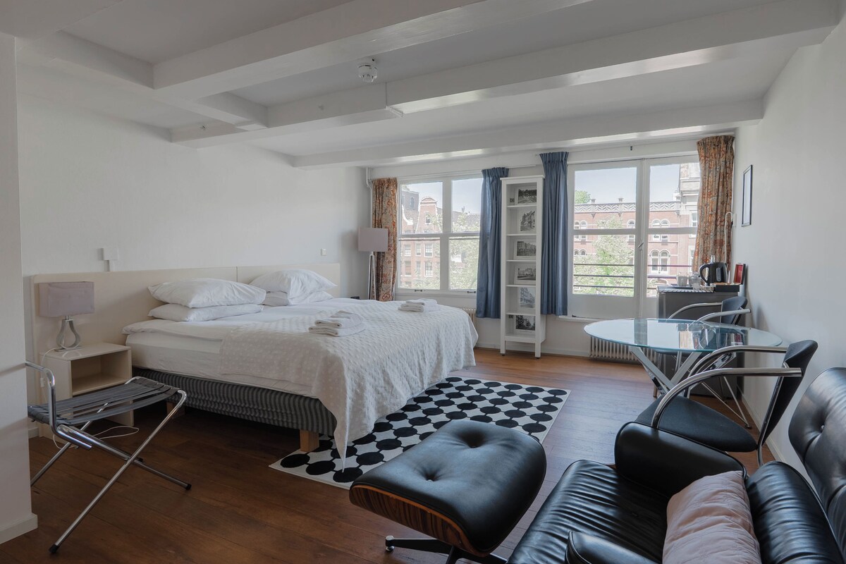 A hotel room with a bed, a desk, a chair, a small table, and a view of the city through large windows.