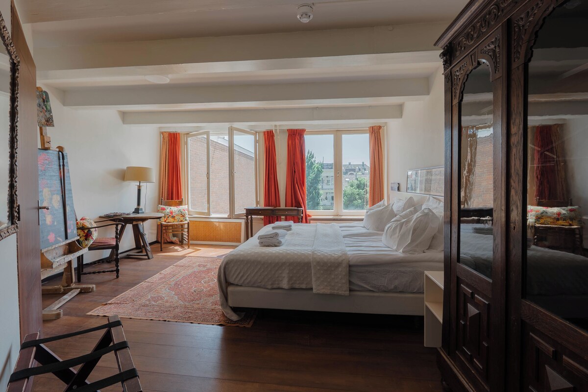 A bedroom with a large bed, a desk with a lamp, a chair, and a view of the outside through a window.