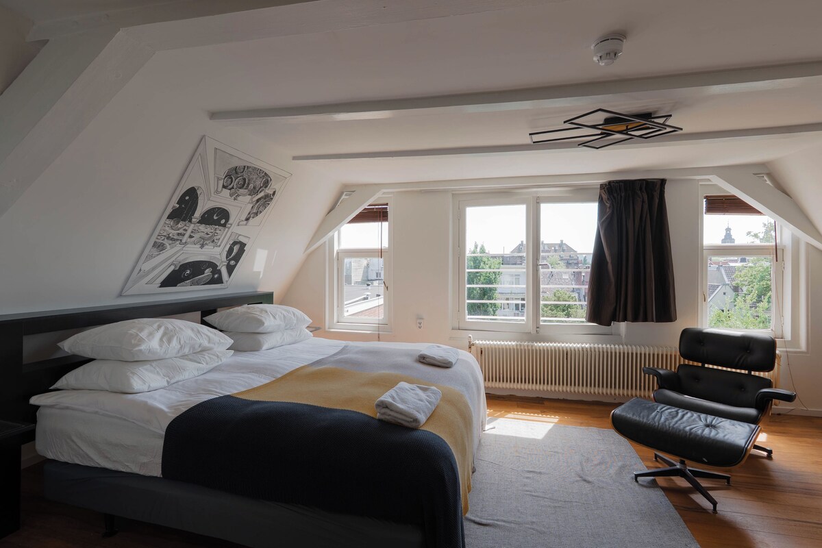 A bedroom with a bed, a chair, a radiator, a window with curtains, and a city view.