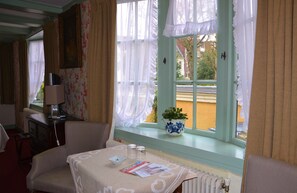 Una habitación de hotel con una mesa de comedor, una silla, una mesita con una lámpara, una ventana con cortinas y una planta en el alféizar.
