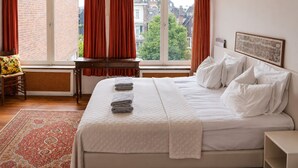 A hotel room with a large bed, a desk, a chair, a window with curtains, and a view of buildings. - Hotel Keizershof (Amsterdam)