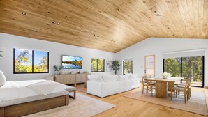 Ein geräumiger Raum mit Holzbalkendecke, einem Bett, einem weißen Sofa, einem Esstisch mit Stühlen und großen Fenstern mit Blick auf Bäume.