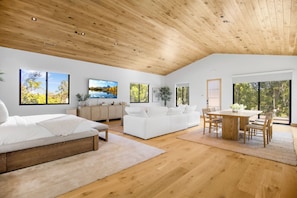 A spacious room with a wooden ceiling, a bed, a white sofa, a dining table with chairs, and large windows offering views of trees. - Lechusa Estate (Malibu)