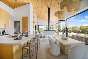 A modern kitchen with a large island, wooden ceiling, and large windows offering a view of the outdoors. - Lechusa Estate (Malibu)