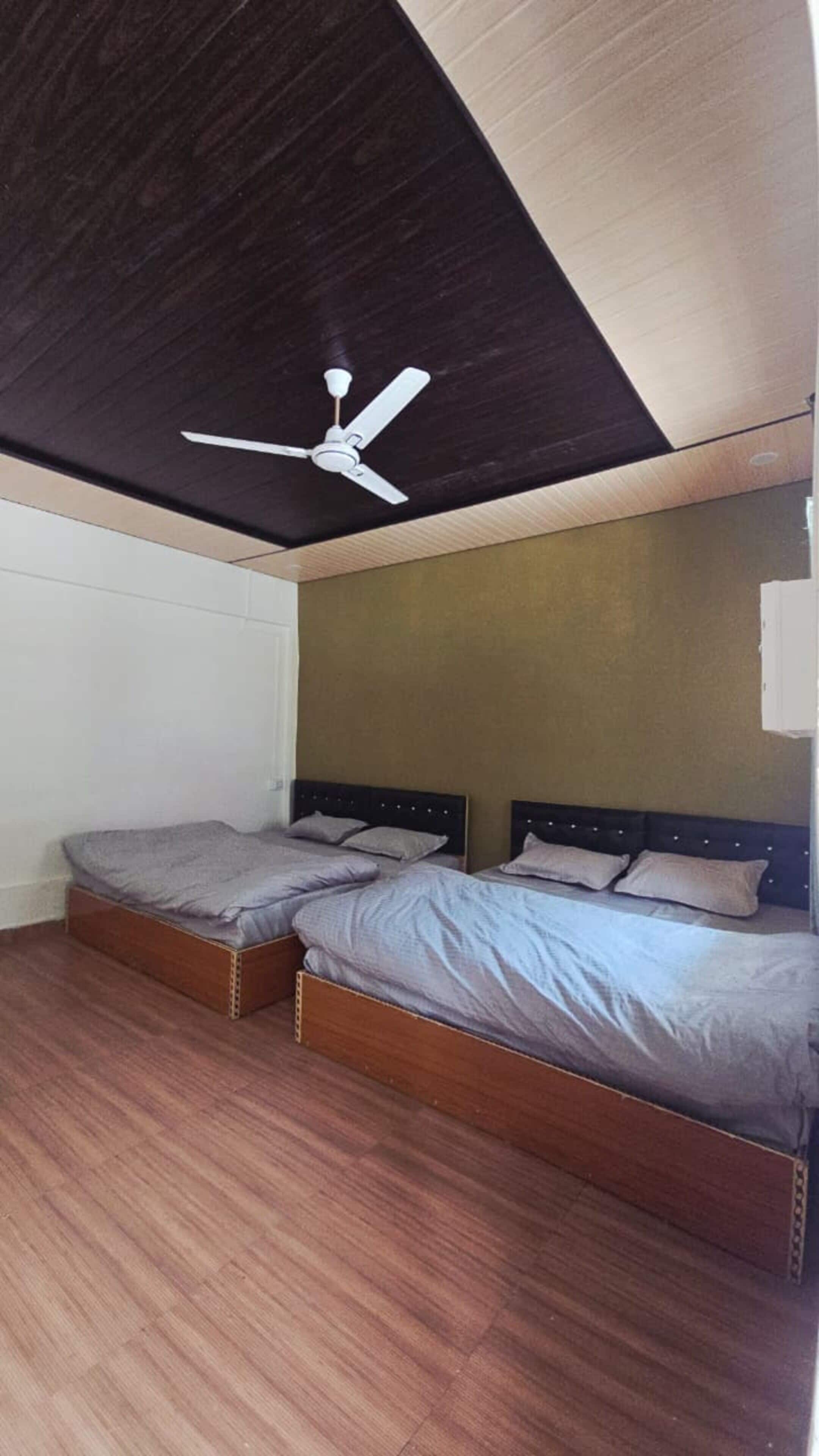 A room with two beds, a ceiling fan, wooden flooring, and a plain wall.